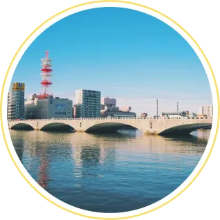 青空のもと、ゆったりと流れる信濃川と、そこに架かるアーチ状の萬代橋を対岸から見渡す新潟市内の風景。