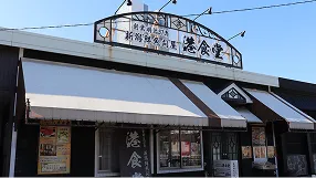 青空を背景にした「新潟鮮魚問屋 港食堂」の店舗外観。黒い壁面の上にレトロなデザインの店名看板が設置されており、白い日よけテントが張られている様子。